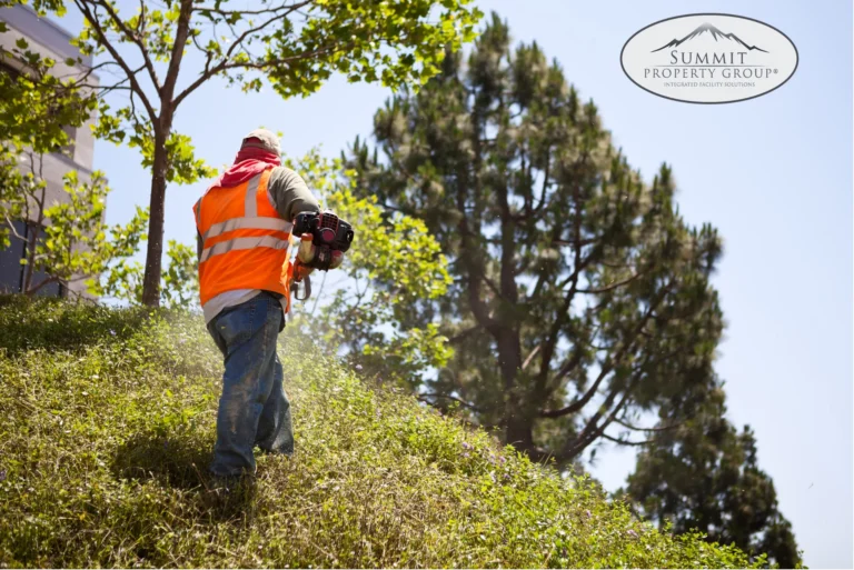 Landscaper in an orange safety vest using a trimmer on a grassy hill surrounded by trees, delivering professional landscaping and property maintenance services in Lethbridge.